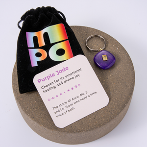 A circular brown presentation tray holding a small purple stone charm, a black velvet pouch with a rainbow 'MPO' logo, and a white card labeled 'Purple Jade' on a white background.
