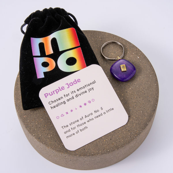 A circular brown presentation tray holding a small purple stone charm, a black velvet pouch with a rainbow 'MPO' logo, and a white card labeled 'Purple Jade' on a white background.