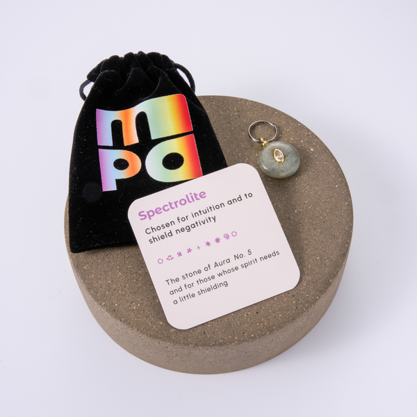 A circular brown presentation tray holding a small green stone charm, a black velvet pouch with a rainbow 'MPO' logo, and a white card labeled 'Spectrolite' on a white background.