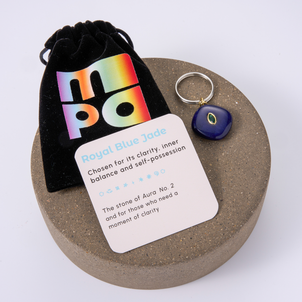 A royal blue jade charm with a silver split ring and black evil eye charm, displayed next to a black velvet pouch with a colorful holographic 'mpa' logo and a white card labeled 'Royal Blue Jade' on a round stone pedestal.