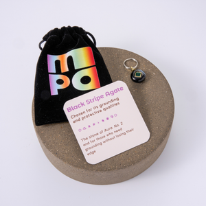 Black Stripe Agate crystal charm with a black velvet pouch and an informational card, displayed on a round wooden tray against a white background.