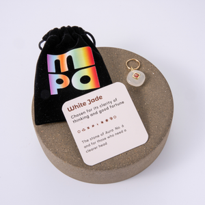 A white jade charm with a gold split ring, displayed next to a black velvet pouch with a colorful holographic 'mpa' logo and a white card labeled 'White Jade' on a round stone pedestal.