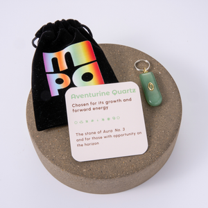Aventurine Quartz charm with a green stone and gold accent, displayed on a round cork coaster alongside a black velvet pouch with a colorful holographic logo and an informational card.