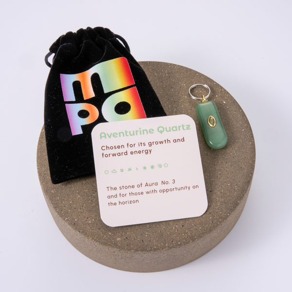 Aventurine Quartz charm with a green stone and gold accent, displayed on a round cork coaster alongside a black velvet pouch with a colorful holographic logo and an informational card.