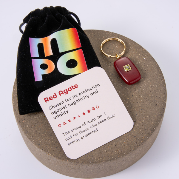 A red agate charm with a gold keyring and a red stone pendant, displayed on a round stone slab next to a black velvet pouch with a rainbow 'mpa' logo and an informational card labeled 'Red Agate'.
