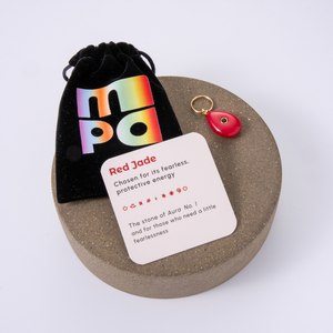 A red jade gemstone charm with a gold keyring, displayed on a round textured gray stone coaster next to a black velvet pouch with a rainbow 'MPA' logo and a white card labeled 'Red Jade'.
