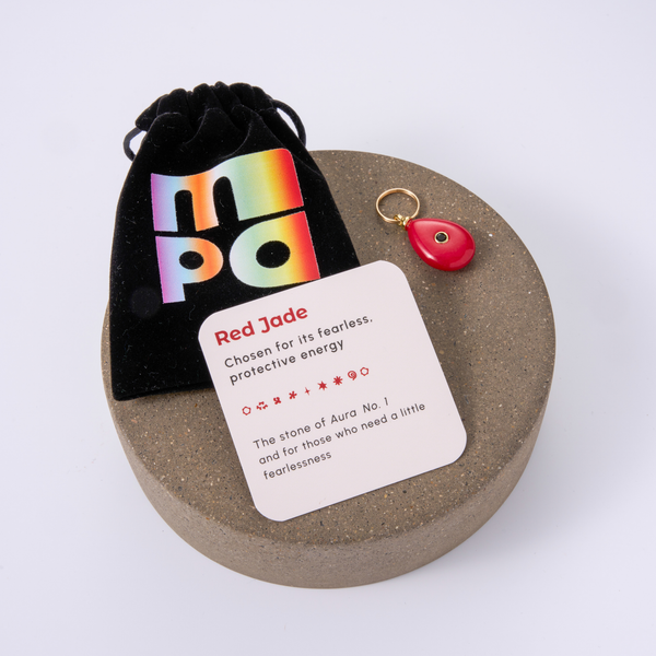 A red jade gemstone charm with a gold keyring, displayed on a round textured gray stone coaster next to a black velvet pouch with a rainbow 'MPA' logo and a white card labeled 'Red Jade'.