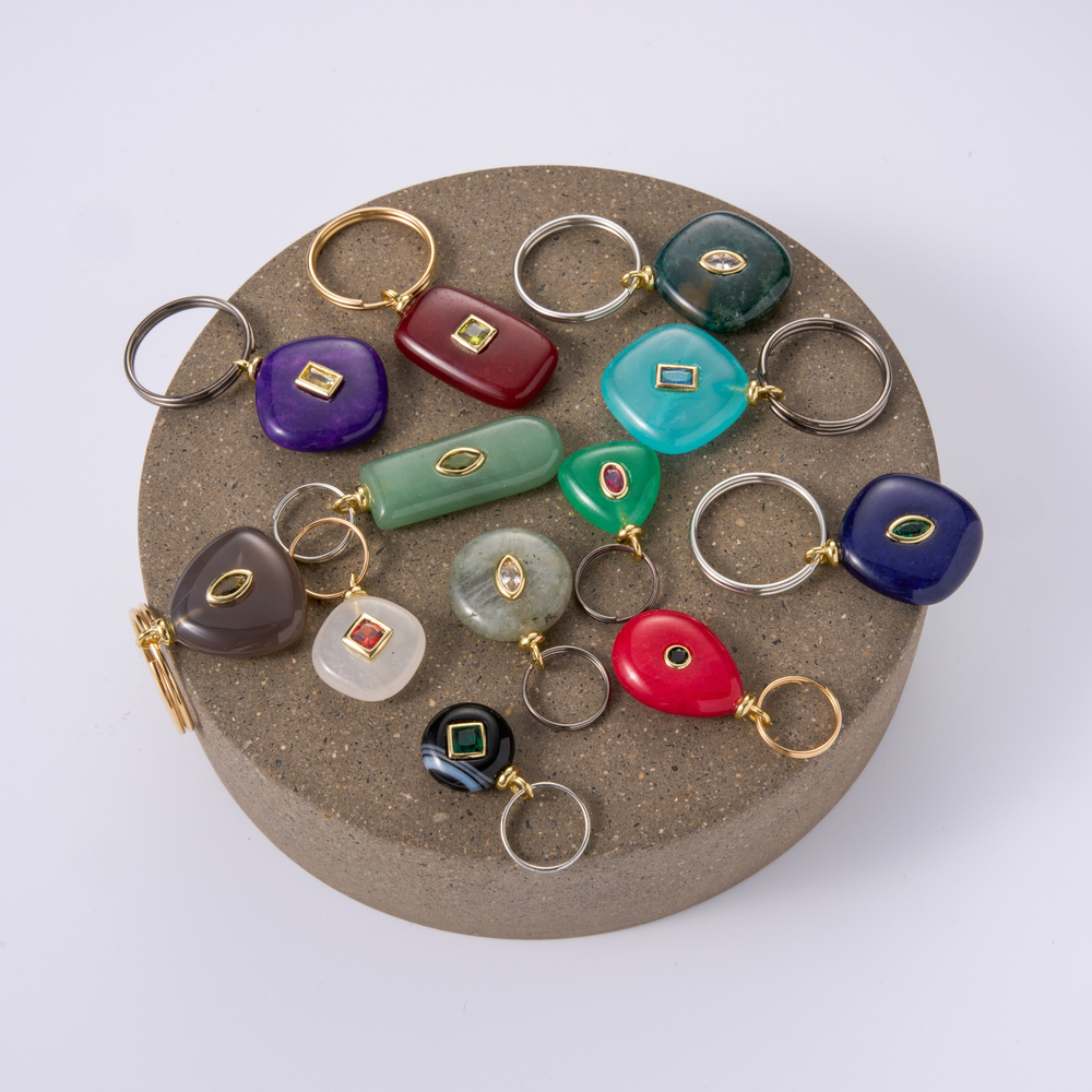 A collection of colorful stone charms with gold-tone rings, displayed on a round textured brown stand against a light gray background.