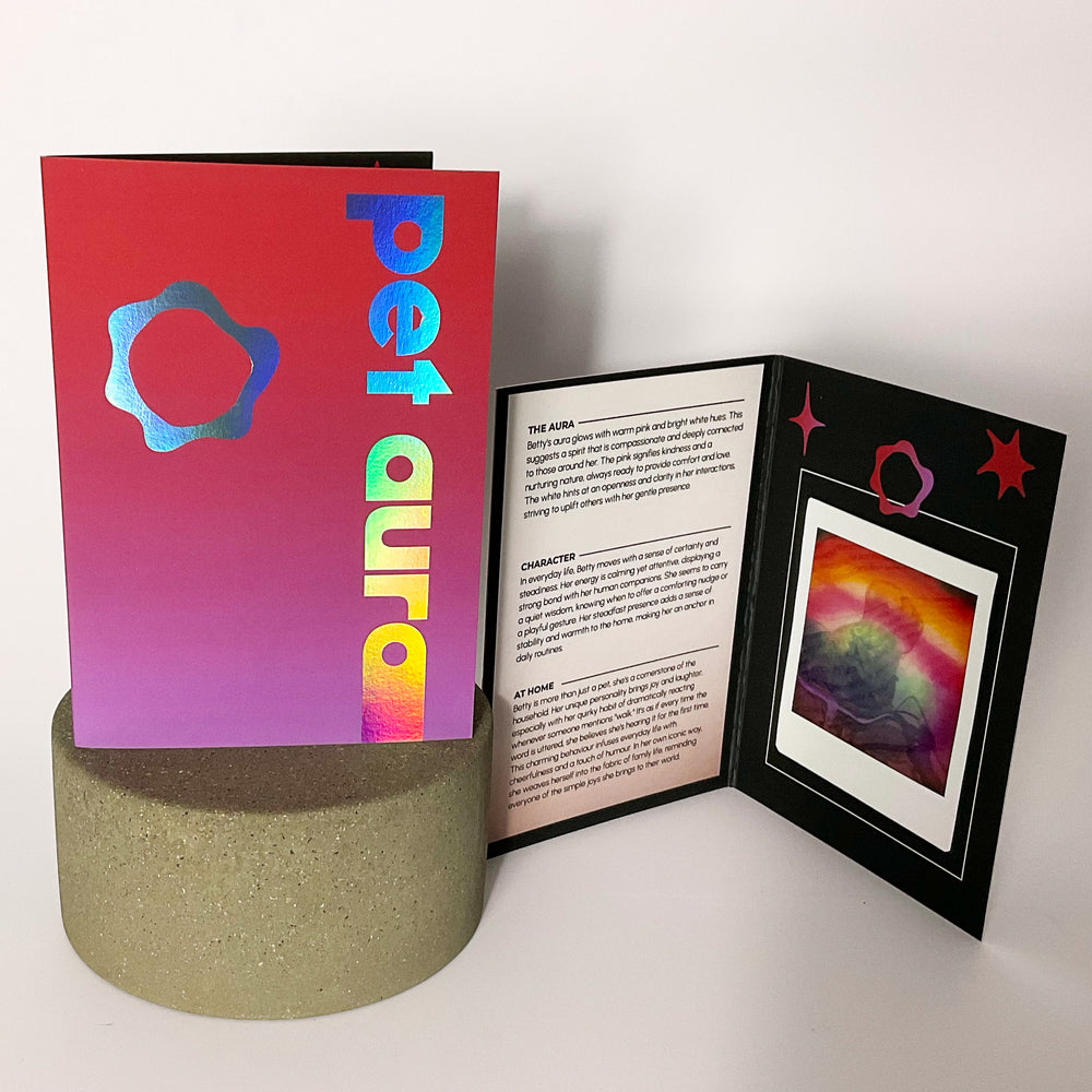 A colorful printed booklet including a pet aura reading displayed on a round textured pedestal against a plain light background. One upright cover is bright pink-to-purple with holographic blue text reading 'pet aura' and a flower-like logo.