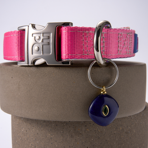 A pink dog collar with gray stitching, a silver metal buckle, and a key ring holding a purple stone charm with a gold accent, displayed on a gray cylindrical pedestal.