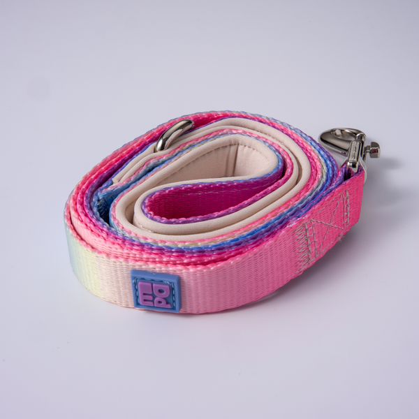 A coiled pink dog leash with a white inner lining, a metal snap hook clasp, and a small square brand patch on a light gray background.