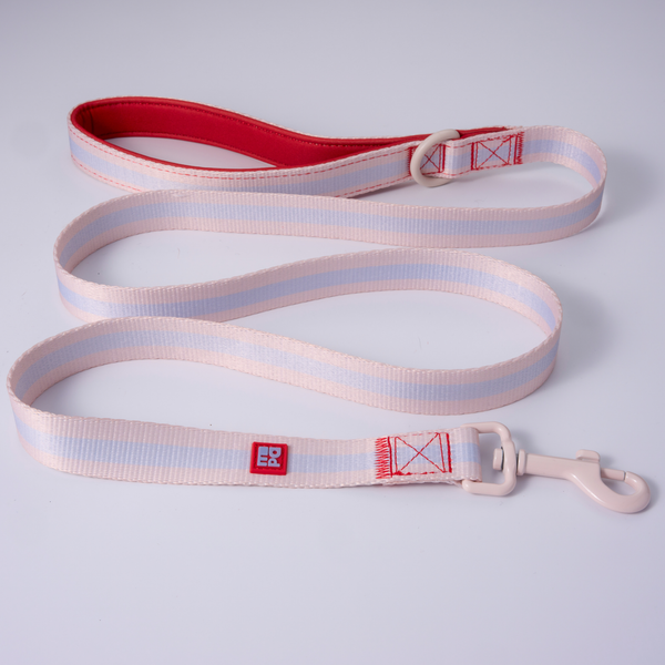 A light pink and white striped fabric dog leash with a red interior lining, beige metal buckle, and a red square logo patch, shown on a light gray background.