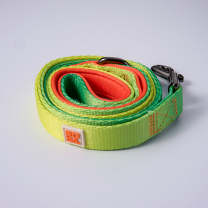 A coiled dog leash made of woven fabric with a gradient of green and yellow, featuring a gray metal clasp and a orange and white brand patch.